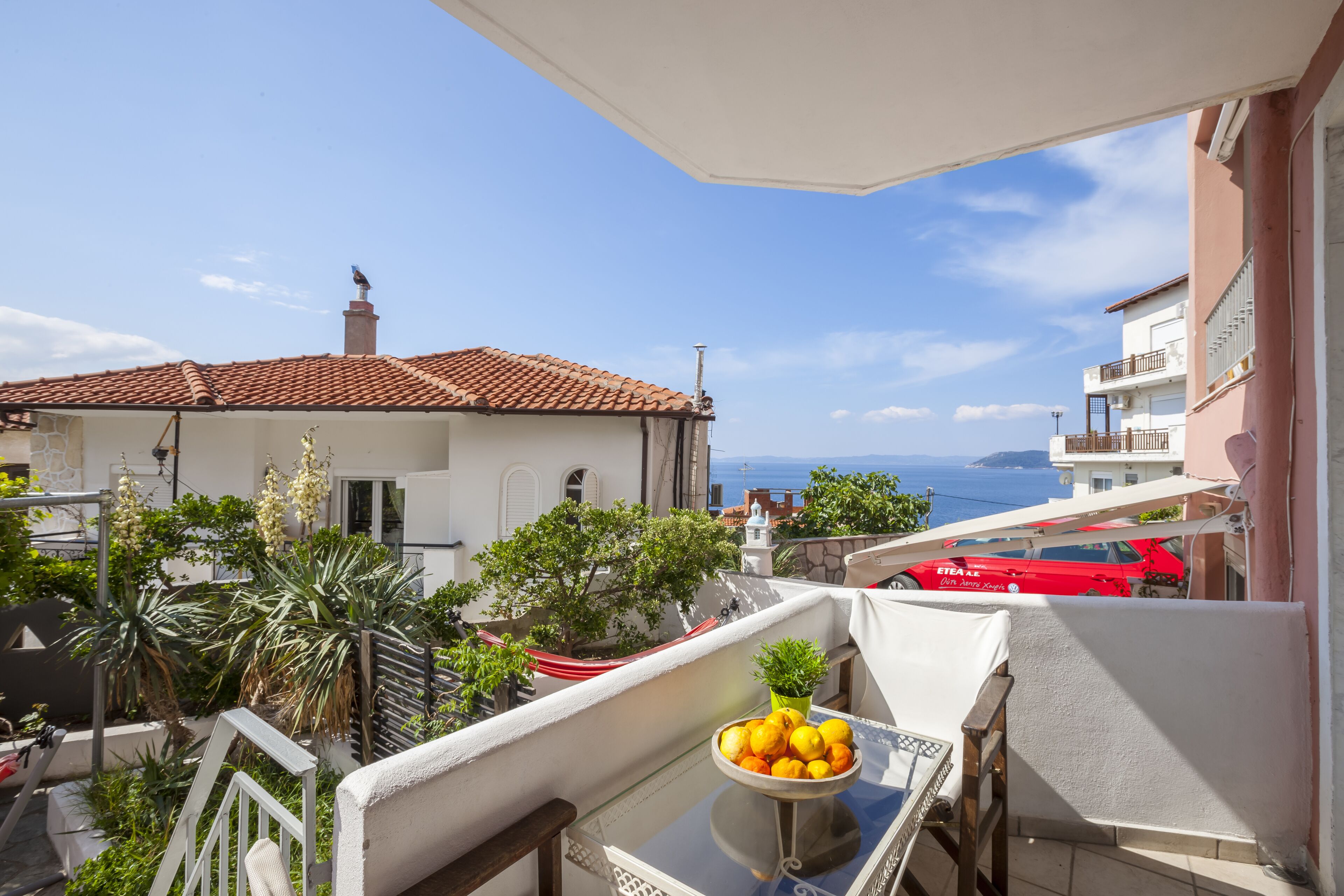 Un balcon avec une table, un panier de fruits et une vue sur la mer et les bâtiments.