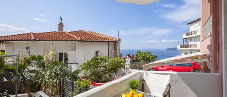 Un balcon avec une table, un panier de fruits et une vue sur la mer et les bâtiments.