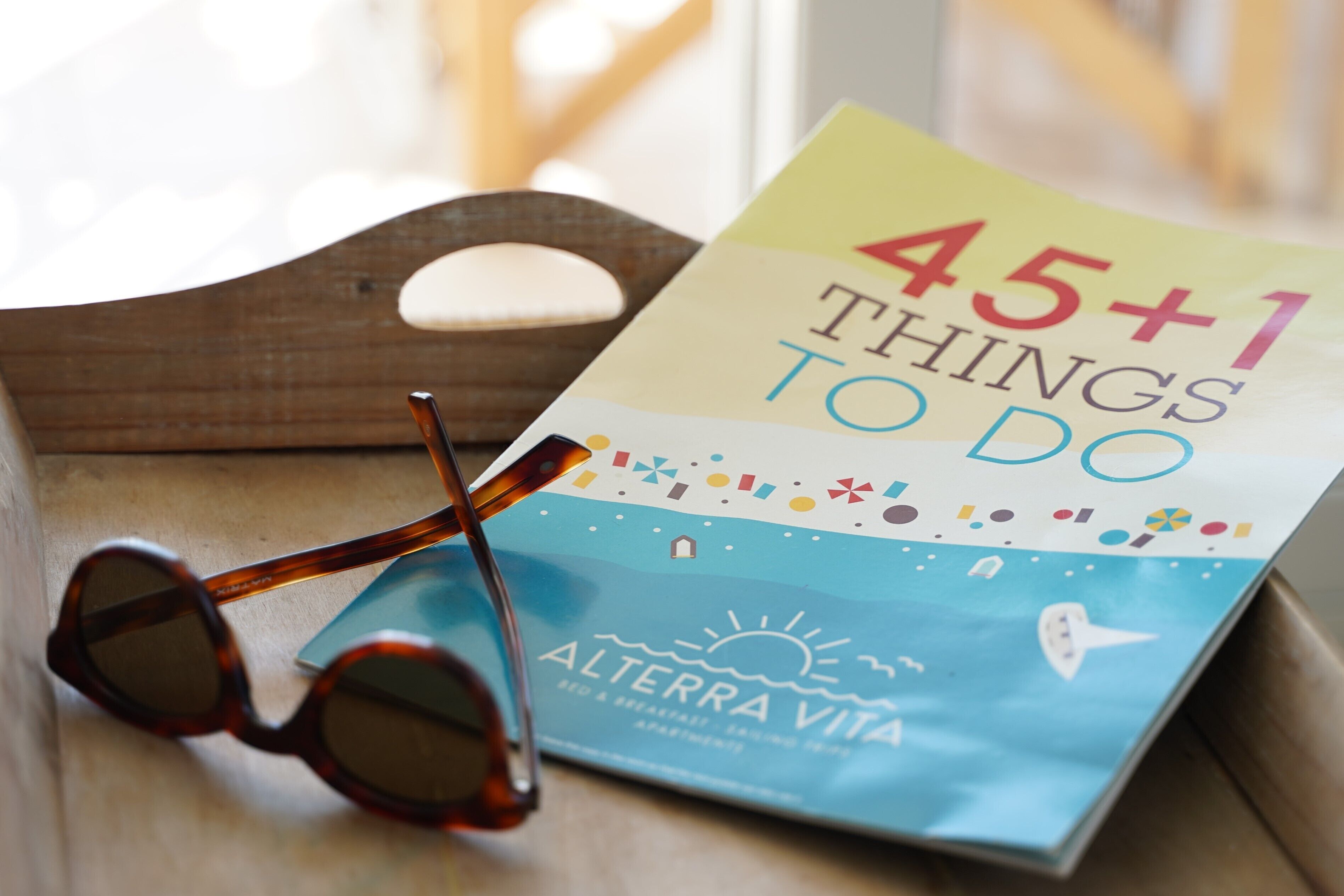 A book titled "45+1 Things to Do" with a beach and sun illustration, placed on a wooden surface with a pair of sunglasses beside it.