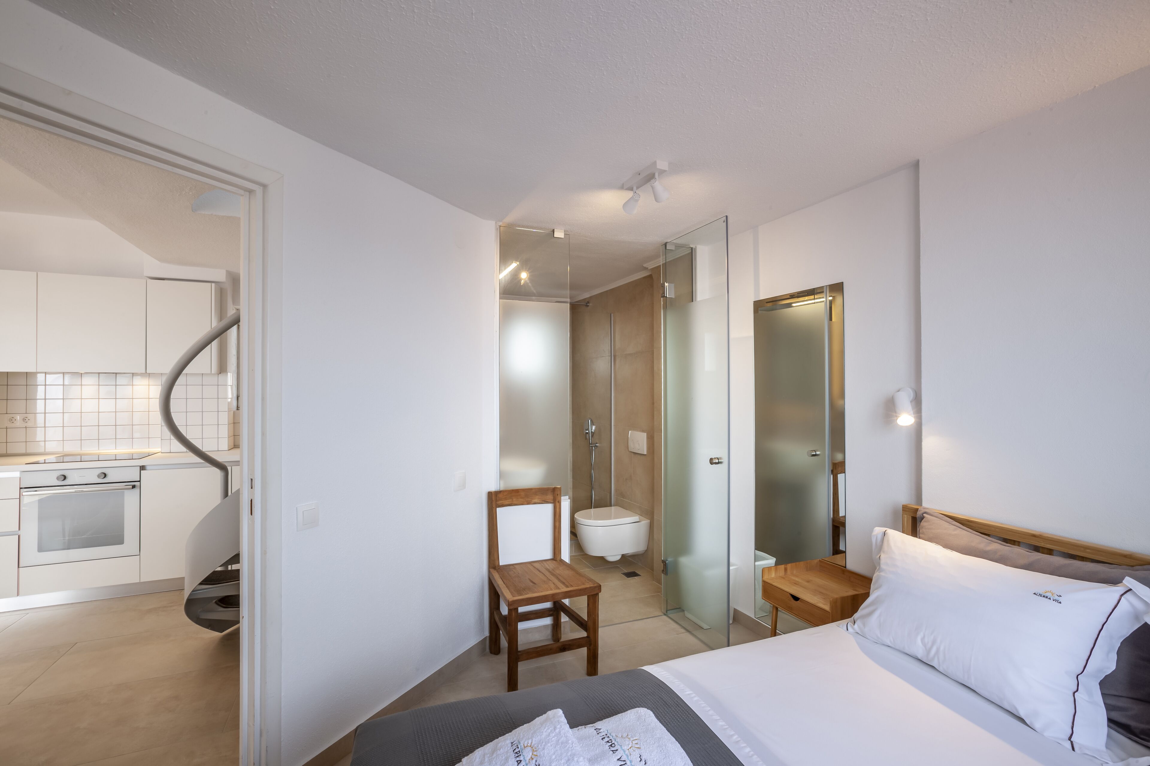 Una habitación de hotel moderna con una cama, una silla de madera, una mesita y un baño visible a través de una puerta de cristal.