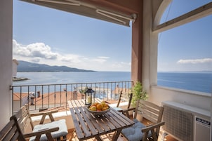 Un balcón con una mesa, sillas, un frutero, un farol y vistas al mar y a las montañas.