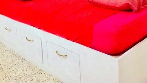 Un lit avec une couvre-lit rouge et deux oreillers, une commode blanche avec des tiroirs et une fenêtre avec un rideau.