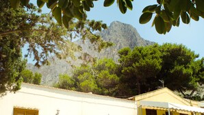 A building with white walls and yellow shutters, a mountain in the background, and greenery in the foreground.