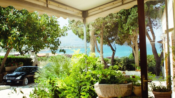 A covered outdoor area with potted plants, a parked car, and a view of the sea.
