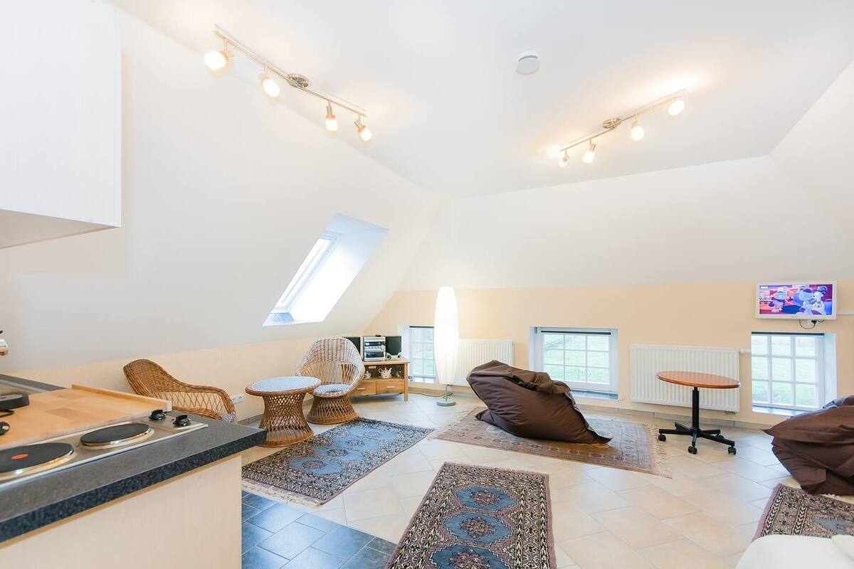 A spacious living area with a kitchen, a dining table, and a sofa. There are large windows providing natural light and a TV mounted on the wall.