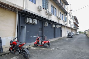 A narrow street with parked scooters and a car, buildings with air conditioning units, and a closed garage door.