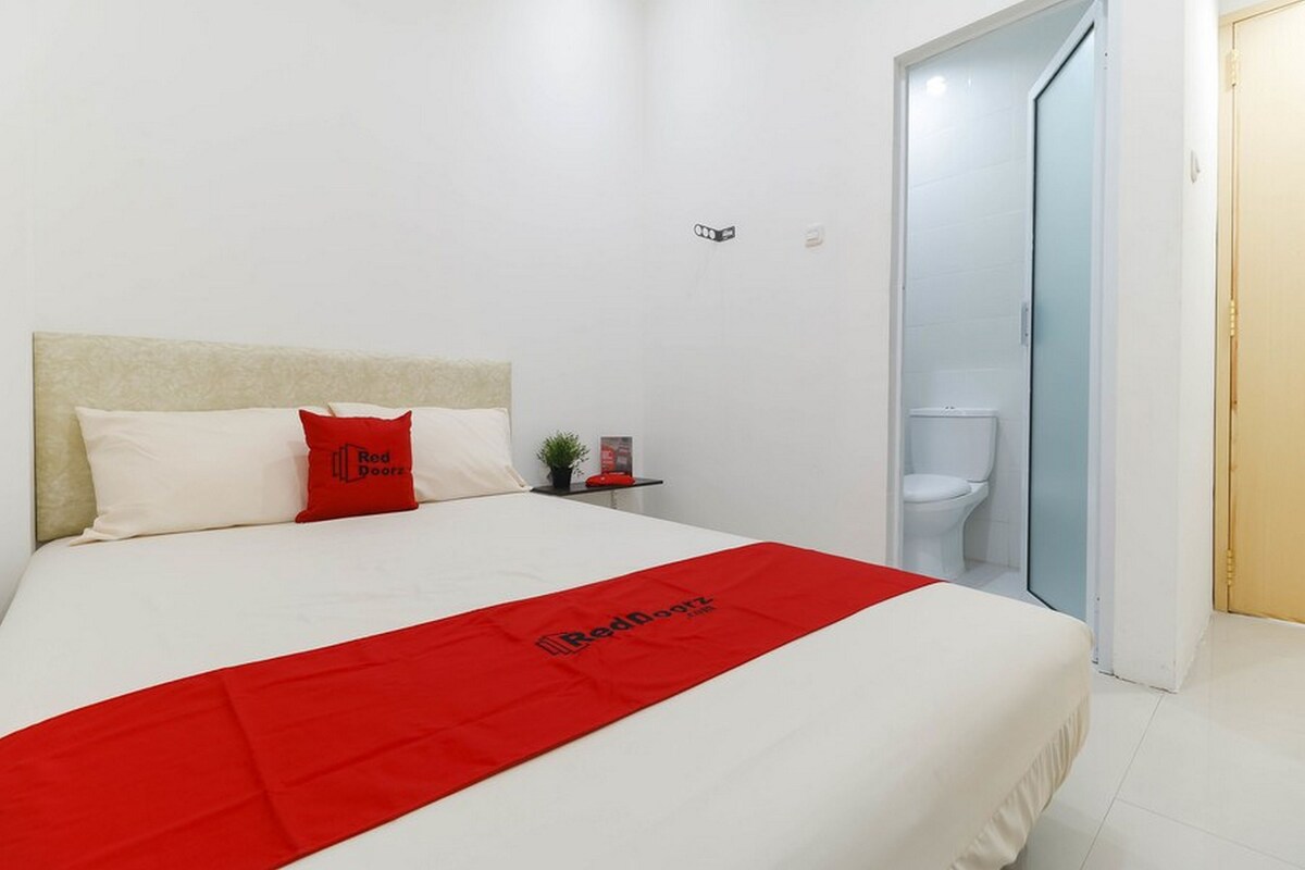 Une chambre d’hôtel avec un lit, une couverture rouge, une petite table de chevet avec une plante, et une porte donnant sur une salle de bain.