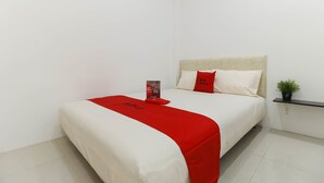Una cama bien hecha con un detalle rojo, una mesita de noche con una planta y un estante en la pared.