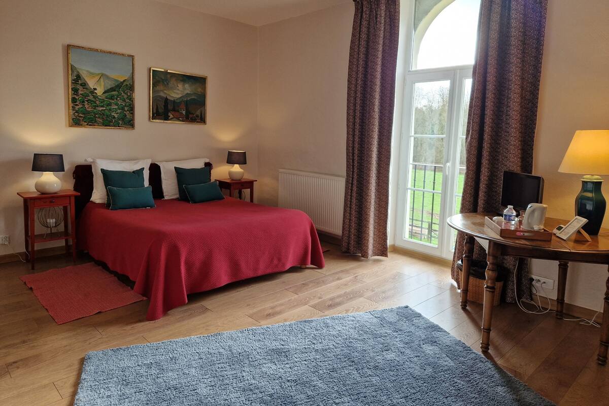 Una habitación de hotel con una cama, dos cuadros, una ventana con cortinas, una mesa con lámpara y una alfombra azul.