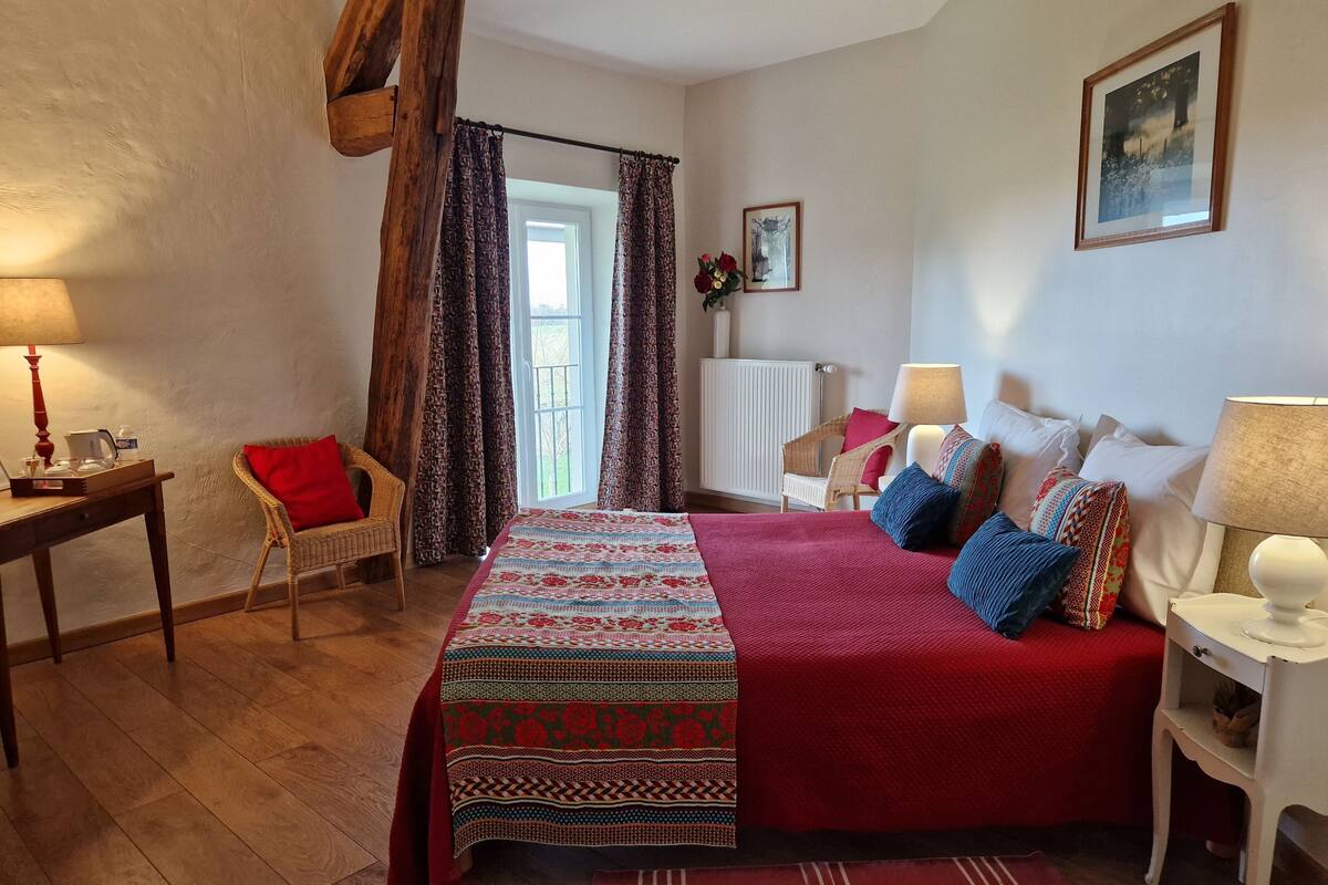Un dormitorio acogedor con una cama grande, una silla de mimbre, una mesita con lámpara y una ventana con cortinas.