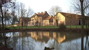 A large, historic stone building with multiple chimneys, reflecting in a calm pond. - Ferme D'Orsonville (Villiers-en-Bière)