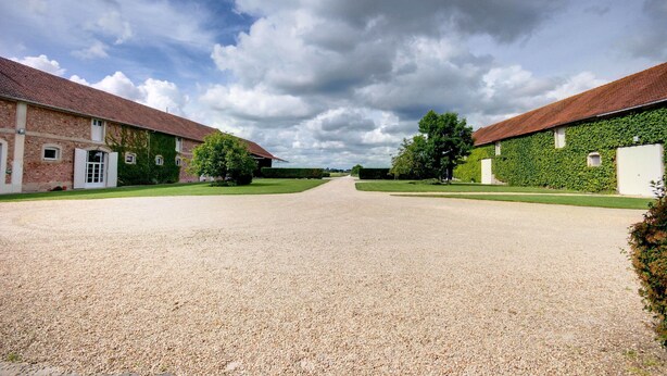 A gravel driveway leading to a building with a red-tiled roof, surrounded by greenery and a clear sky. - Ferme D'Orsonville (Villiers-en-Bière)