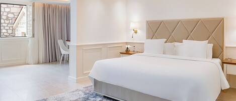 A modern hotel room with a large bed, a desk, a chair, and a view of a stone wall through a window.