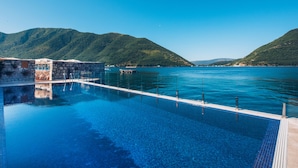 A clear blue pool with a stone wall and a wooden deck, surrounded by lush green hills and a calm sea. - Heritage Grand Perast (Perast)