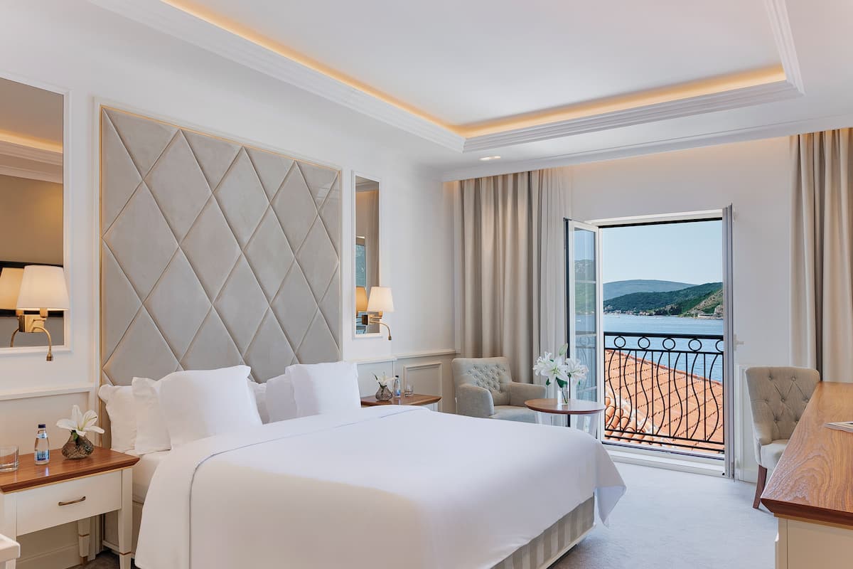 A hotel room with a large bed, a seating area with two chairs, a small table, and a view of the sea through a balcony.