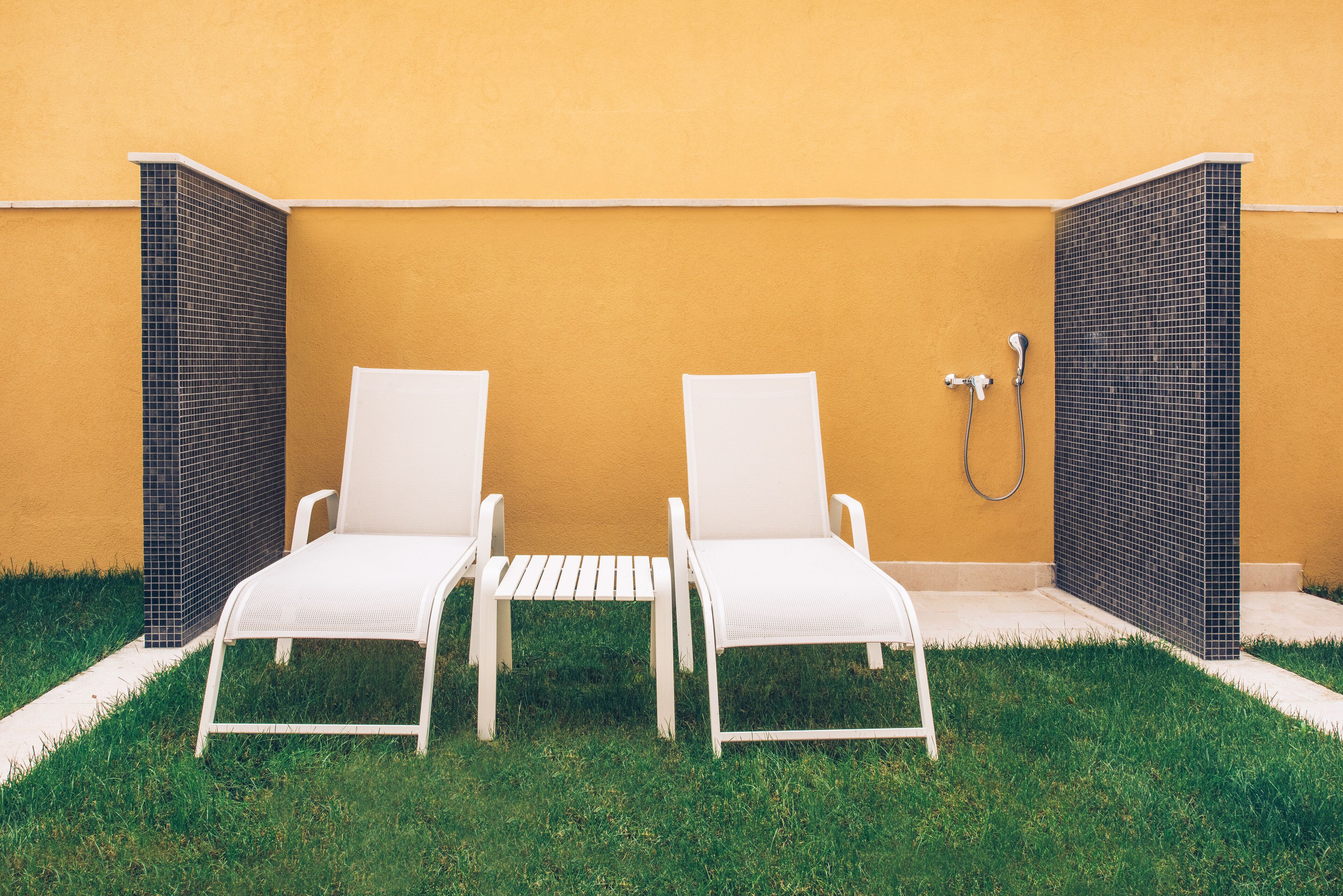 Two white lounge chairs with a small table between them, positioned against a yellow wall with a tiled shower area to the right.