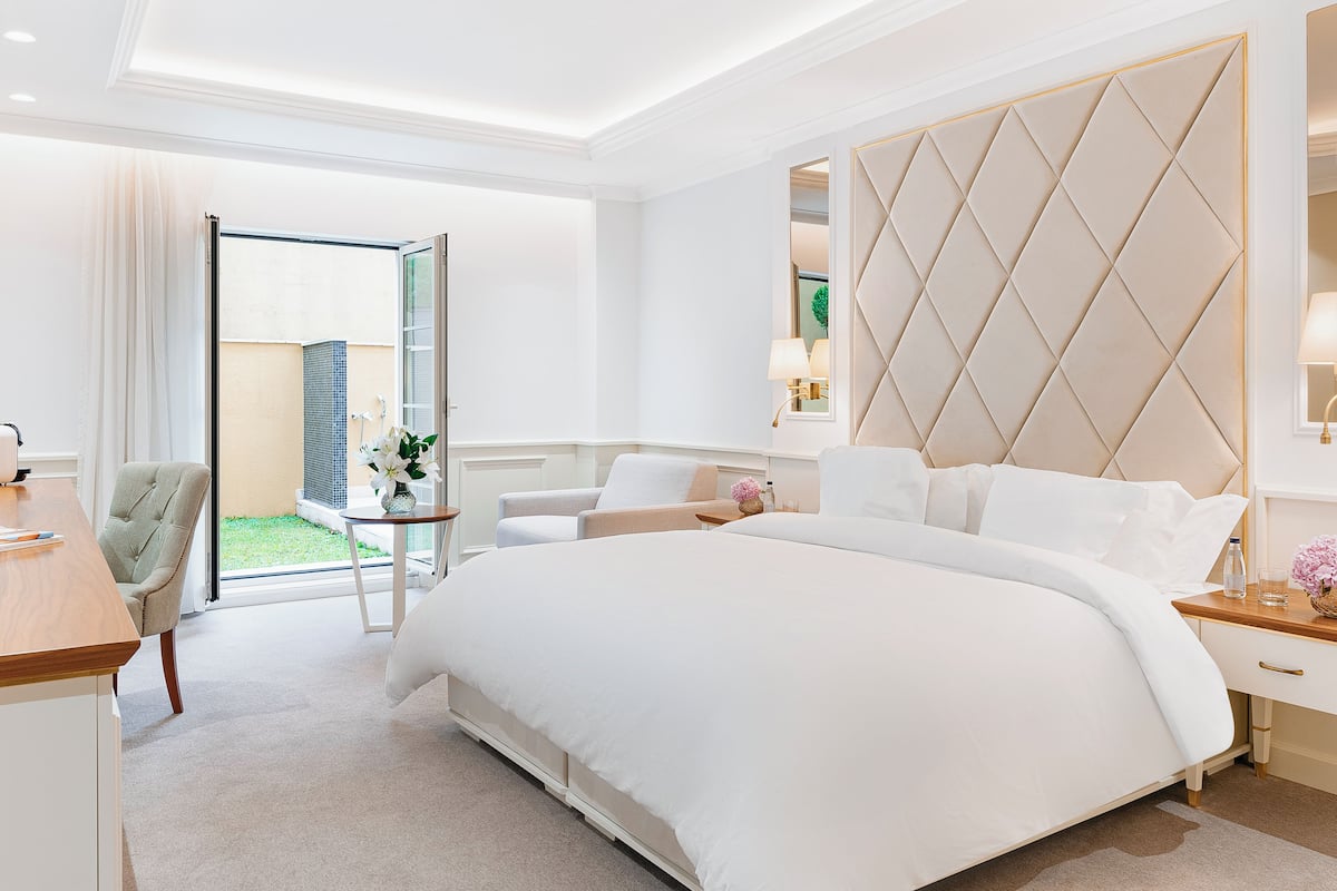 A modern hotel room with a large bed, a desk, a chair, a small table with a lamp, and a view of the outside.