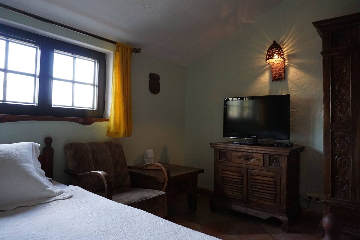 A room with a bed, a wooden chair, a small table, a television on a wooden cabinet, and a window with a yellow curtain.