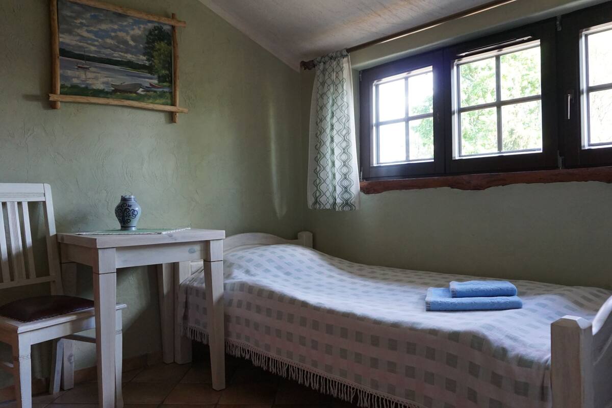 A small, simple room with a bed, a chair, a table, and a painting on the wall.