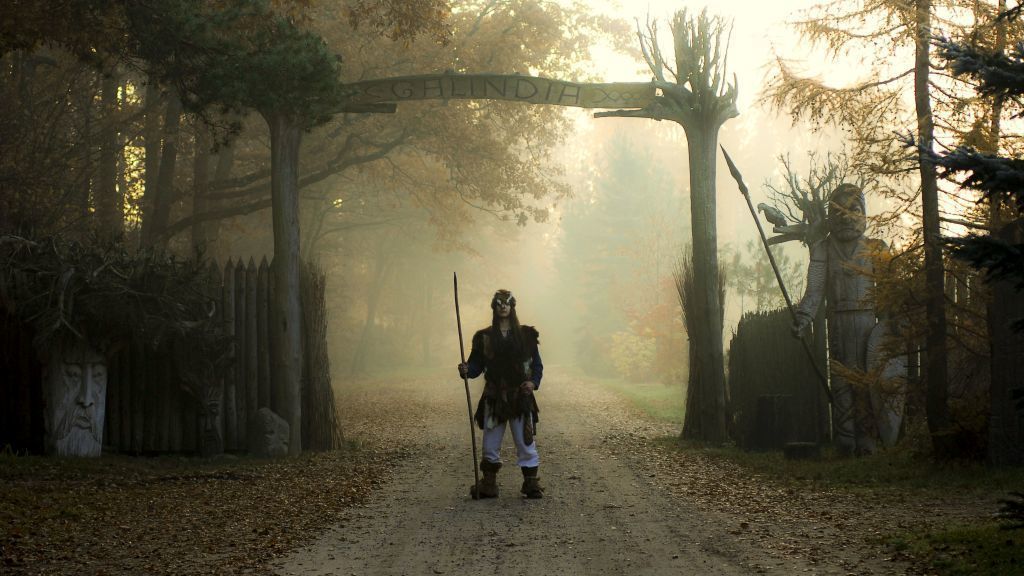 A person standing on a dirt path in a forested area with a large archway in the background.