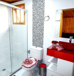 Un baño con cabina de ducha de vidrio, un inodoro con asiento decorado con motivos florales, un lavabo rojo con un jarrón de flores rojas y un espejo sobre el lavabo.