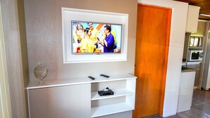 Un salon moderne avec un téléviseur à écran plat fixé sur une console blanche, une porte en bois et un coin cuisine à l’arrière-plan.