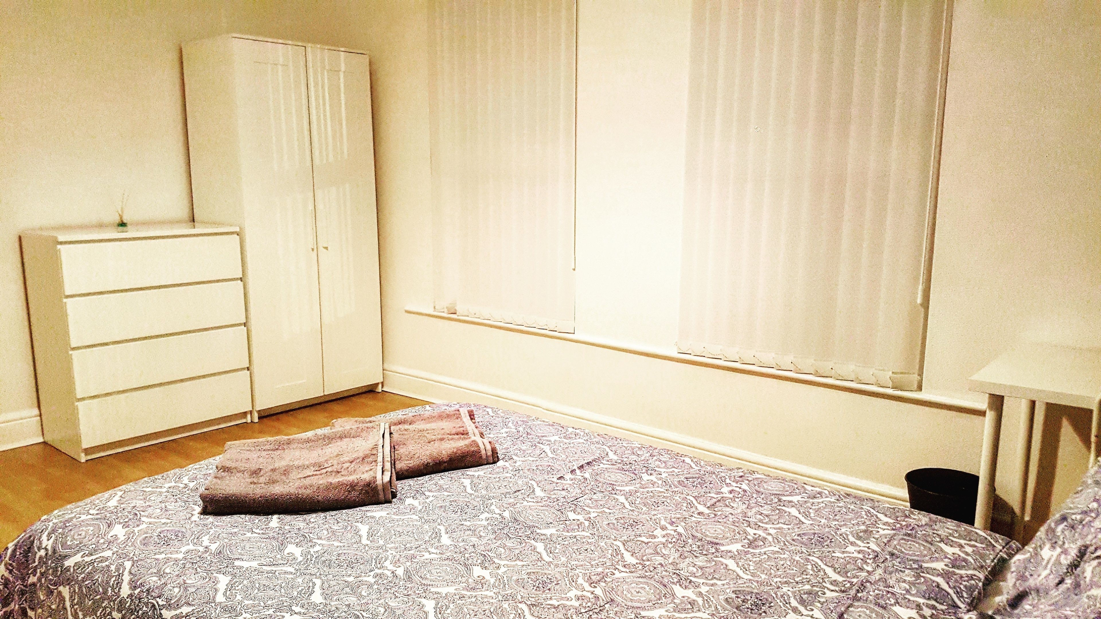 A bedroom with a bed, a wardrobe, a window with blinds, and a nightstand.