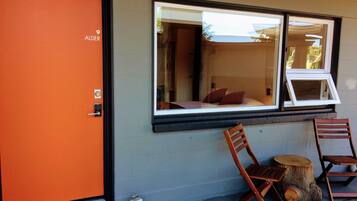Quarto de hotel com porta laranja, uma cama com almofadas e duas cadeiras de madeira na parte externa.