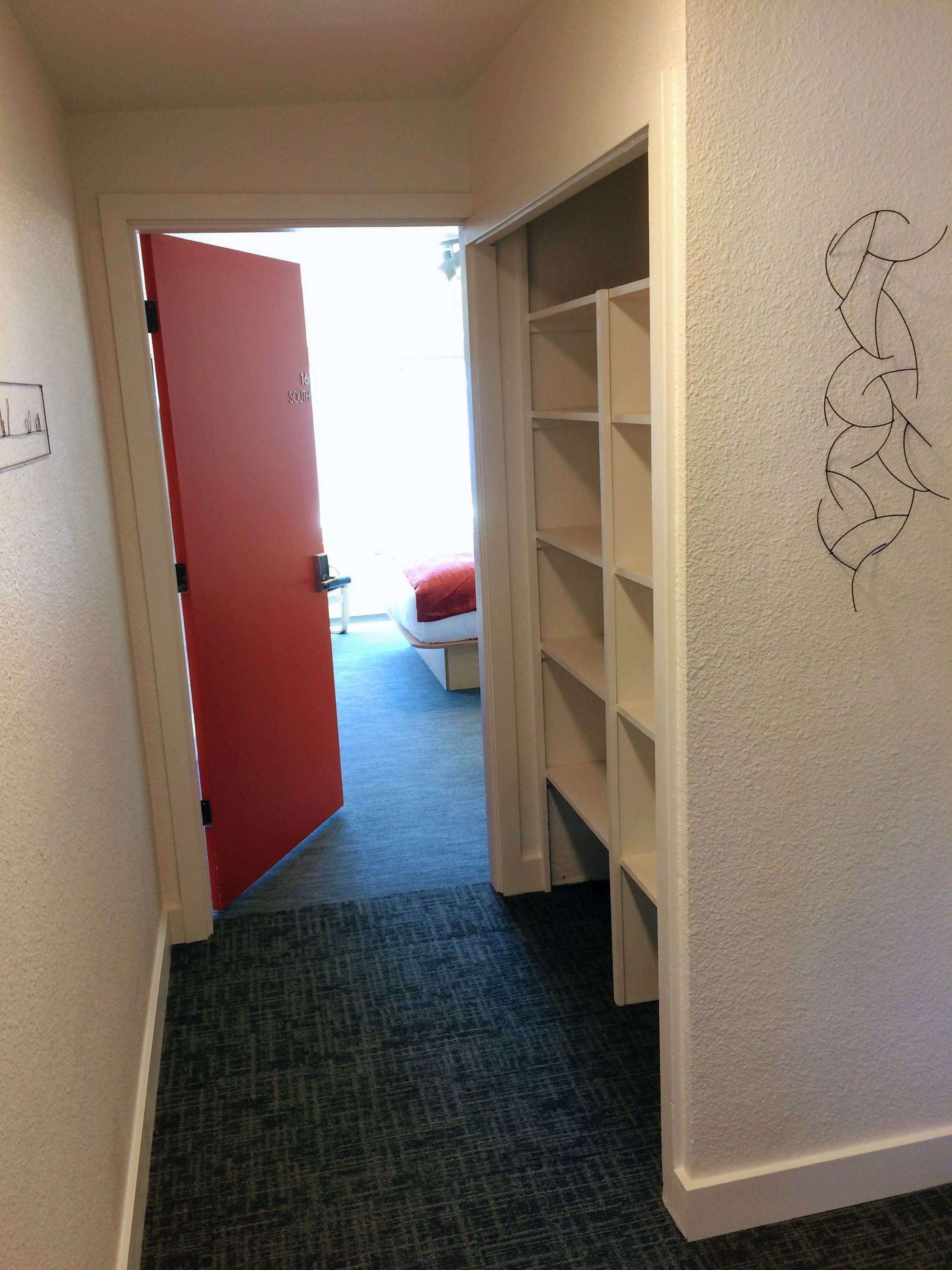 A hallway with a red door, a white shelving unit, and a carpeted floor.