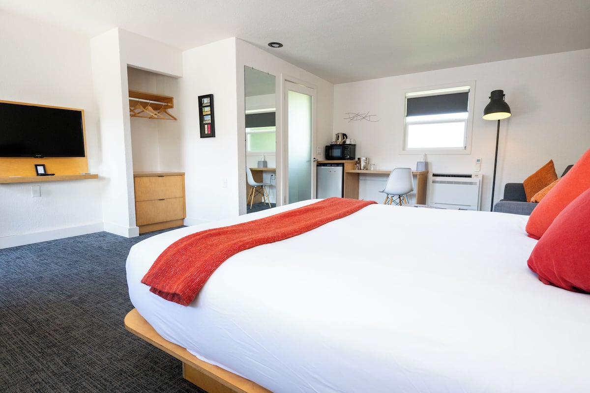 A hotel room with a large bed, a TV mounted on the wall, a desk with a chair, a microwave, and a small kitchenette.