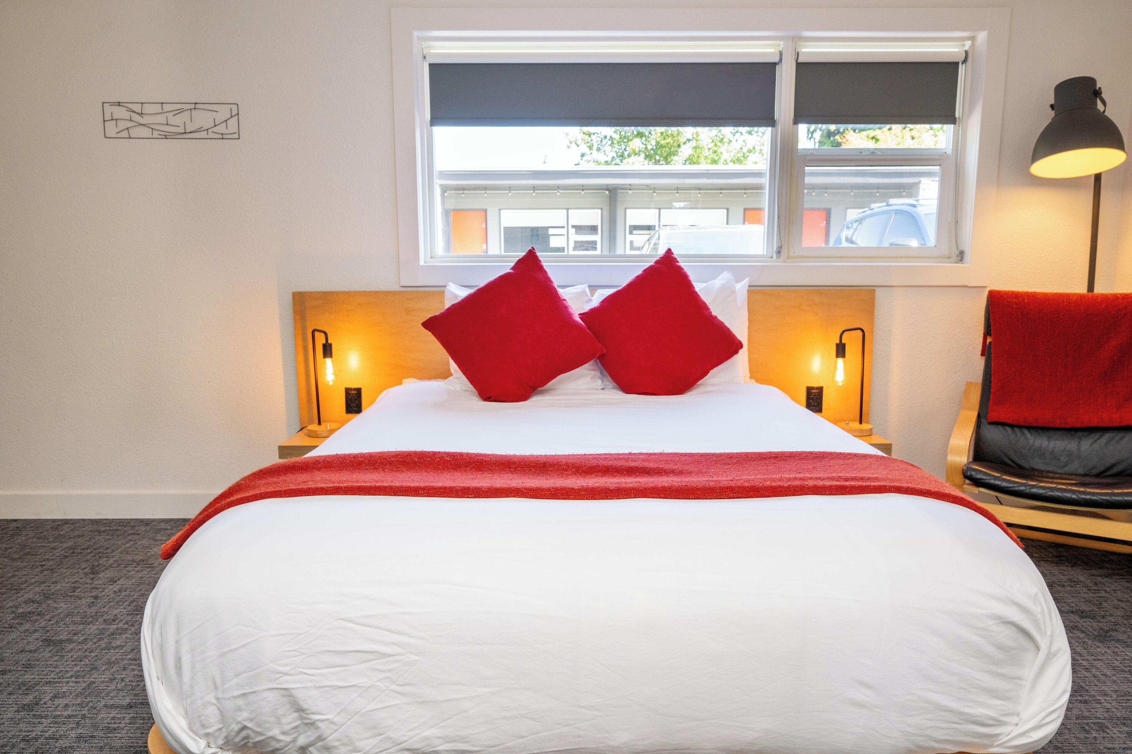 Una cama bien hecha con ropa de cama blanca y almohadas rojas, dos lámparas de noche, una ventana con persianas y una toalla roja sobre una silla.