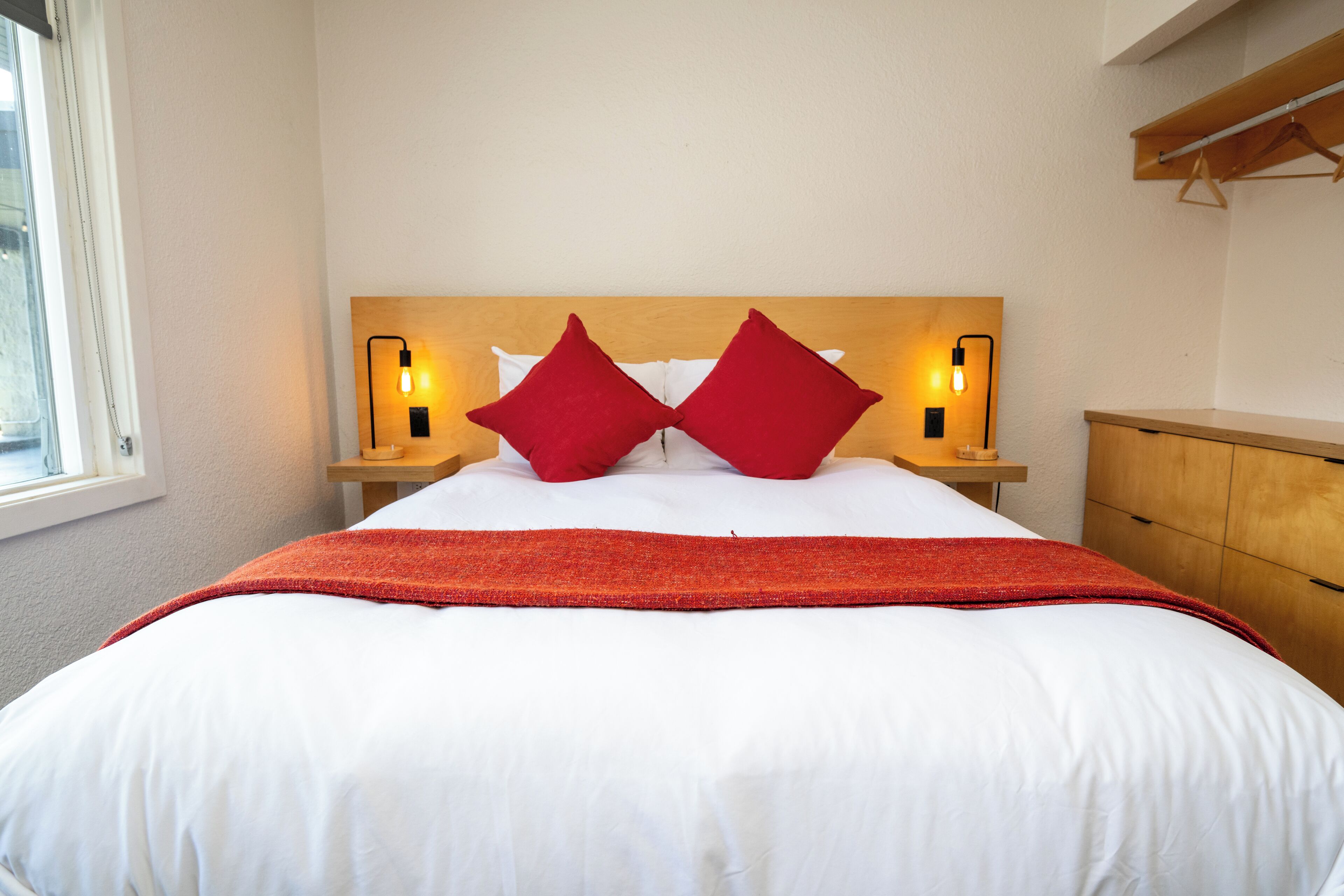 A neatly made bed with white linens and two red pillows, flanked by bedside tables with lamps.