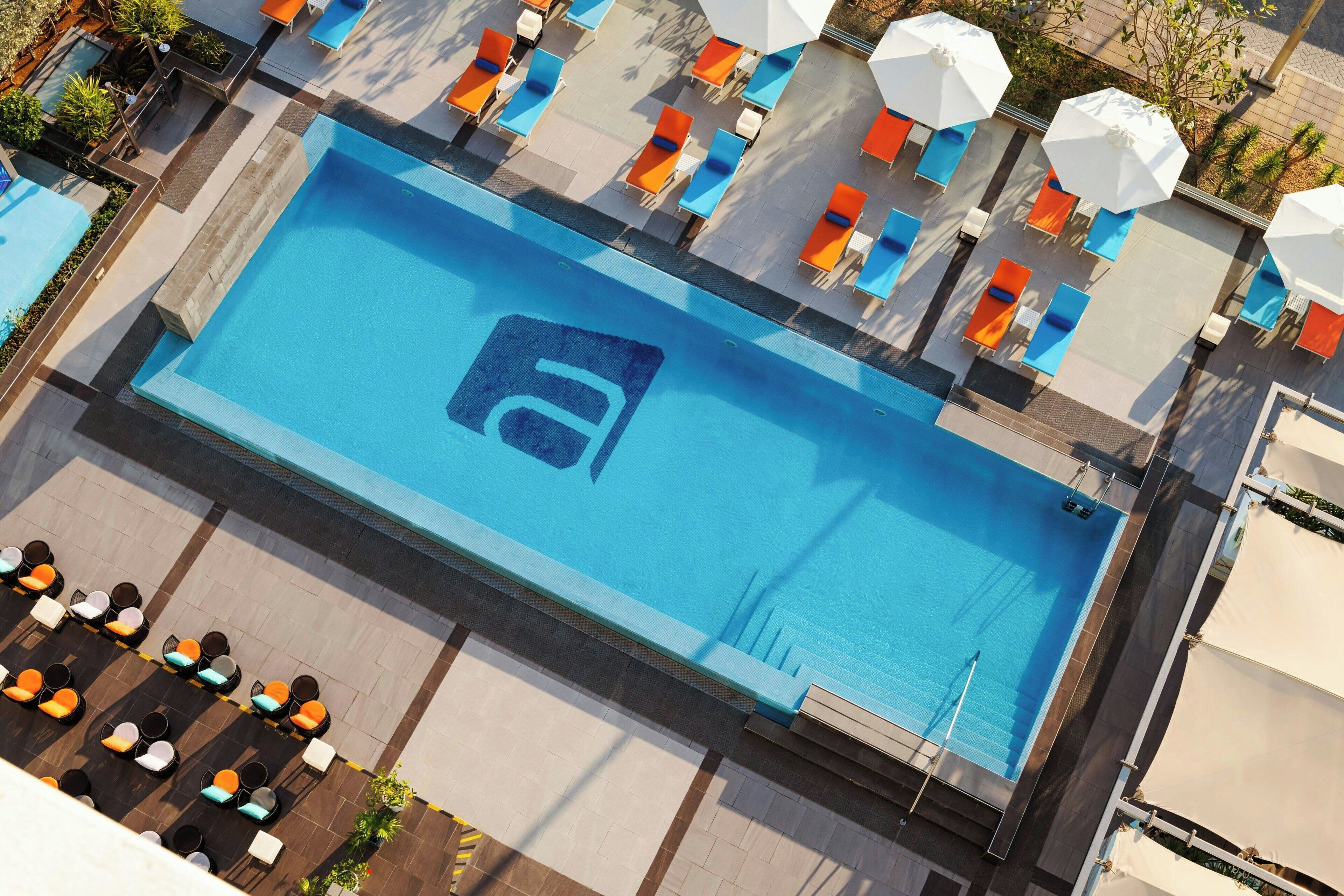 A rooftop pool with a blue logo, orange and white lounge chairs, and umbrellas.