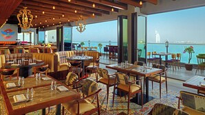 A restaurant with tables and chairs arranged for dining, large windows offering a view of the sea and a prominent building. - Aloft by Marriott Palm Jumeirah (Dubai)