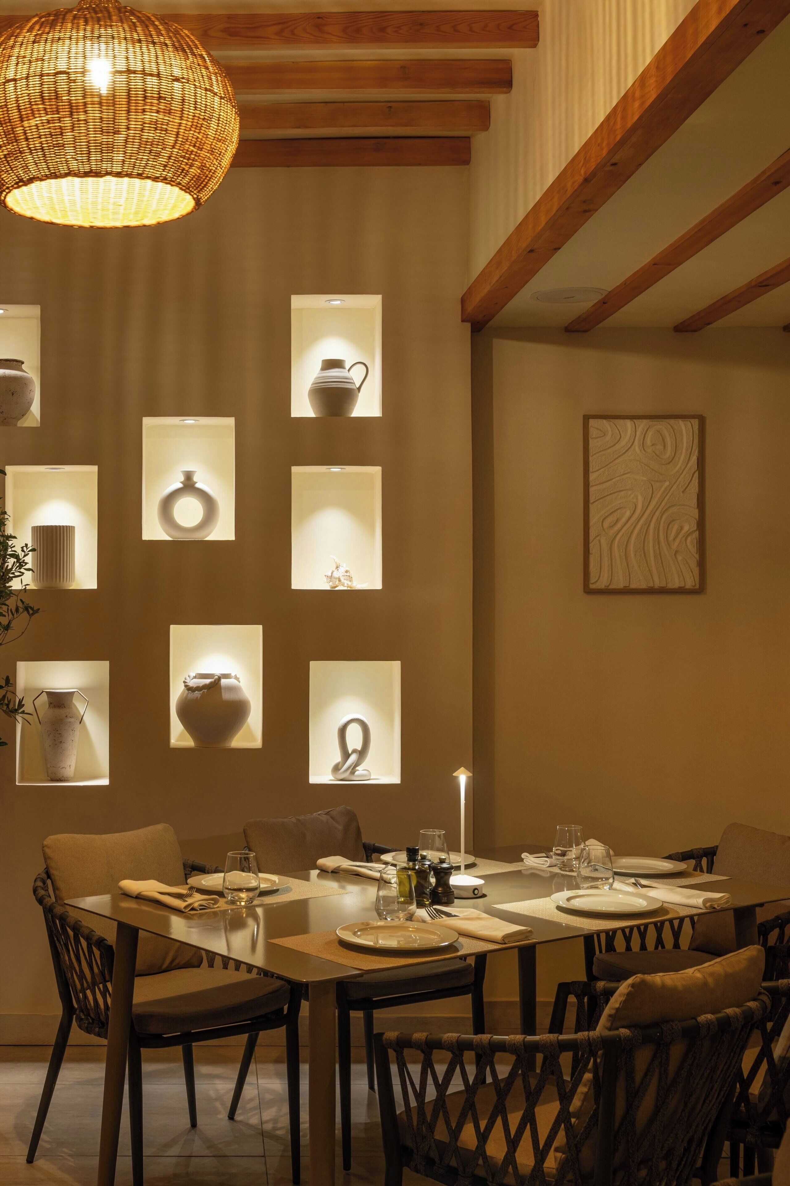A dining area with a large table set for four, surrounded by wicker chairs. There are illuminated shelves displaying various ceramic items.