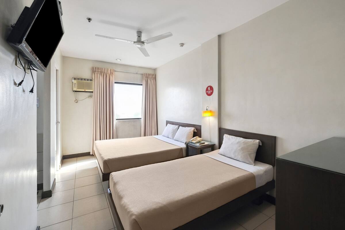 A hotel room with two beds, a ceiling fan, an air conditioning unit, a TV mounted on the wall, and a window with curtains.