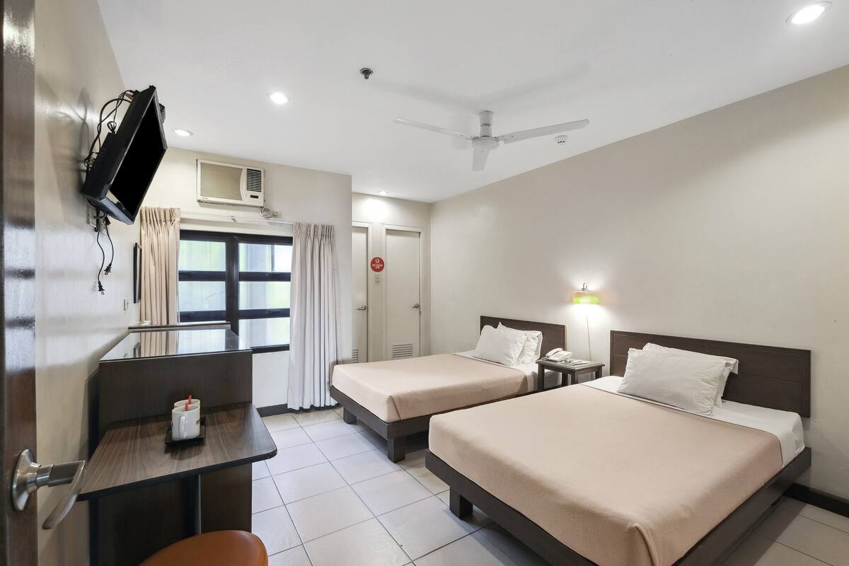 A hotel room with two beds, a desk, a TV mounted on the wall, and a ceiling fan.