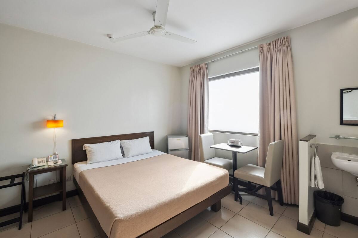 A hotel room with a bed, bedside table, lamp, desk, chair, and a window with curtains.