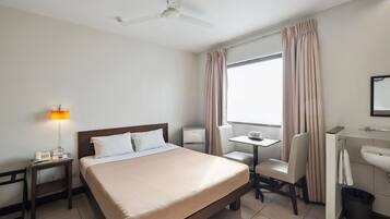 Una habitación de hotel con cama, mesita de noche, lámpara, escritorio, silla y ventana con cortinas.
