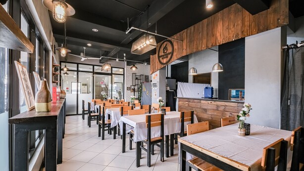 A restaurant interior with wooden tables and chairs, a kitchen area, and pendant lighting.