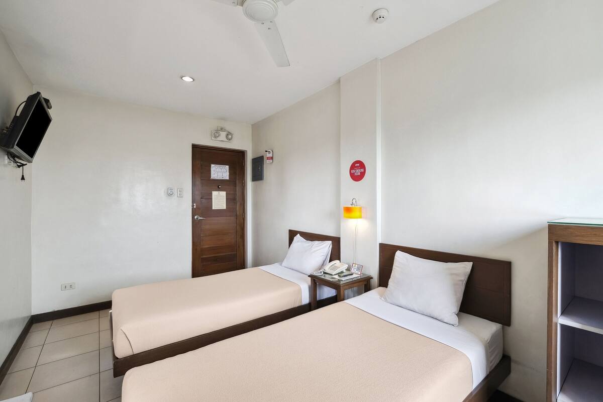 A hotel room with two single beds, a TV mounted on the wall, a ceiling fan, and a door with a sign.