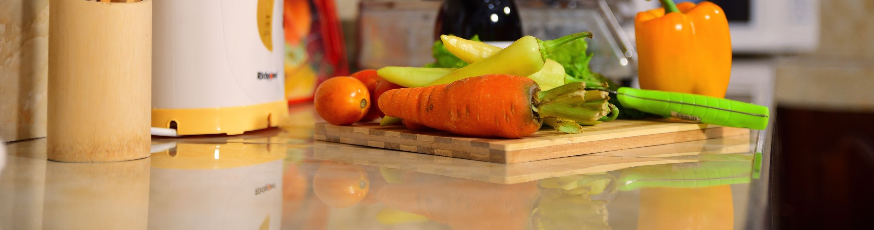 Un plan de travail de cuisine avec un robot culinaire, une planche à découper et divers légumes, notamment des carottes, des poivrons et des oignons.