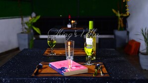 A table with a book, a bottle of wine, and glasses, set against a green backdrop with potted plants. - Kings Villa (Matara)