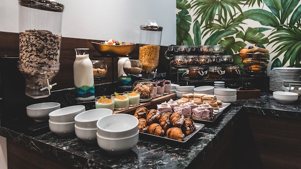 A buffet with various food items and beverages, including a coffee dispenser, a bowl of cereal, and a tray of pastries.