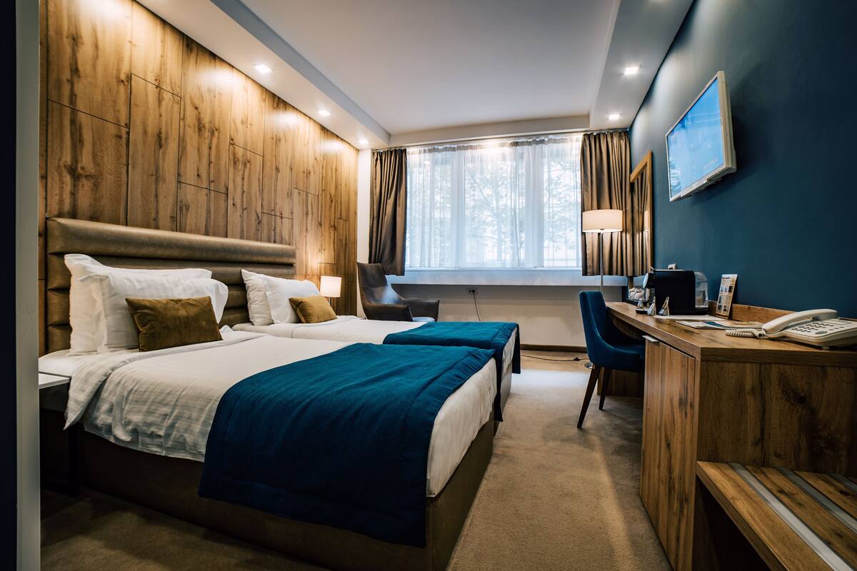 A hotel room with two beds, a desk, a chair, a TV, and a window with curtains.