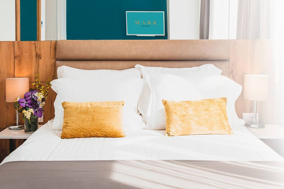 A neatly made bed with white and gold pillows, a wooden headboard, and a teal wall with a sign above.