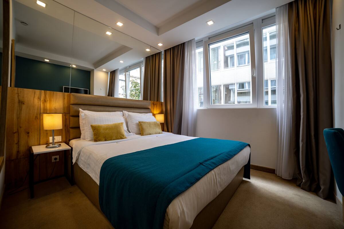 A hotel room with a large bed, a bedside table with a lamp, a view of the city through the window, and a chair by the window.