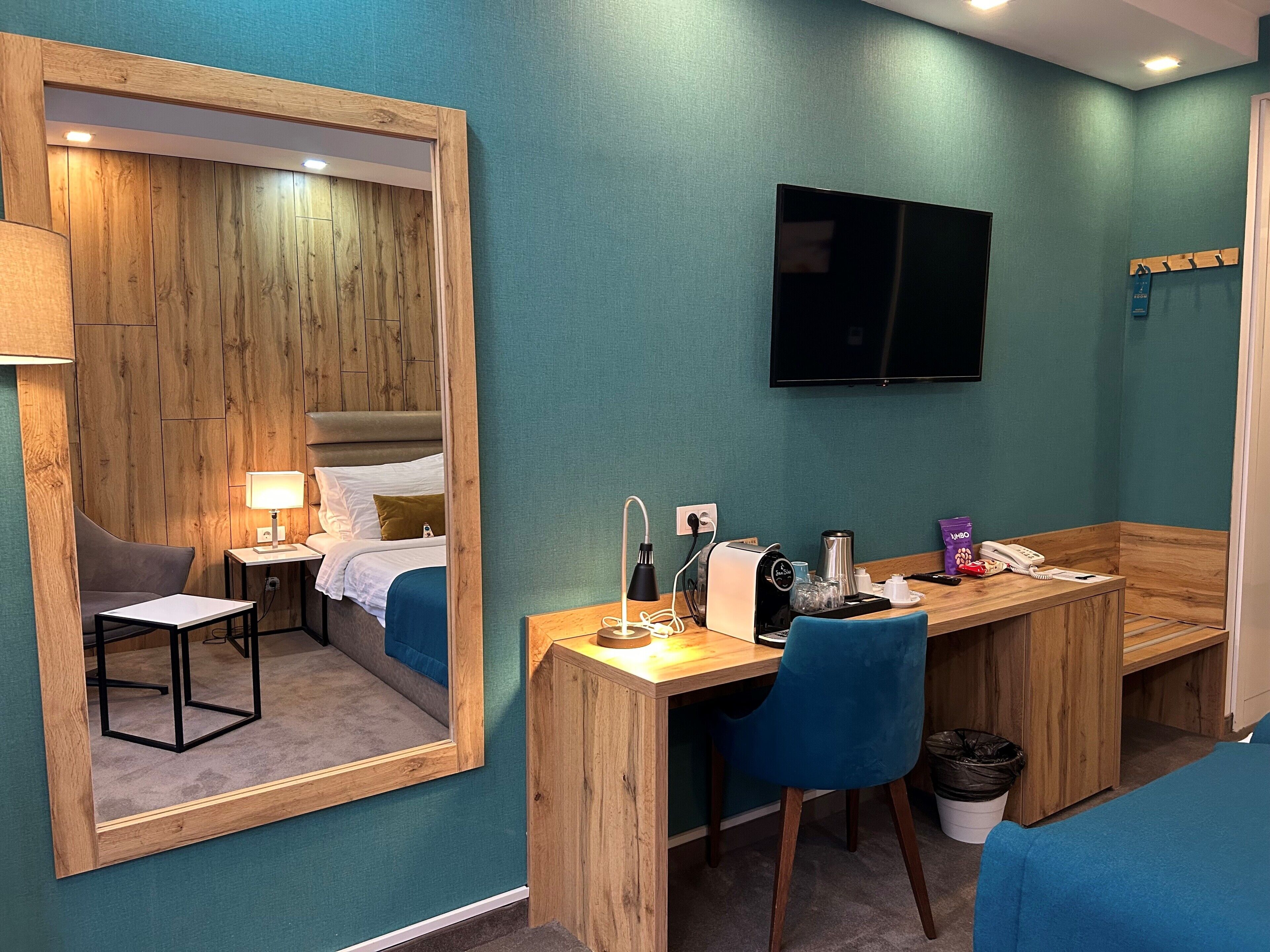 A hotel room with a wooden desk, a chair, a bed with a headboard, a TV mounted on the wall, and a mirror.