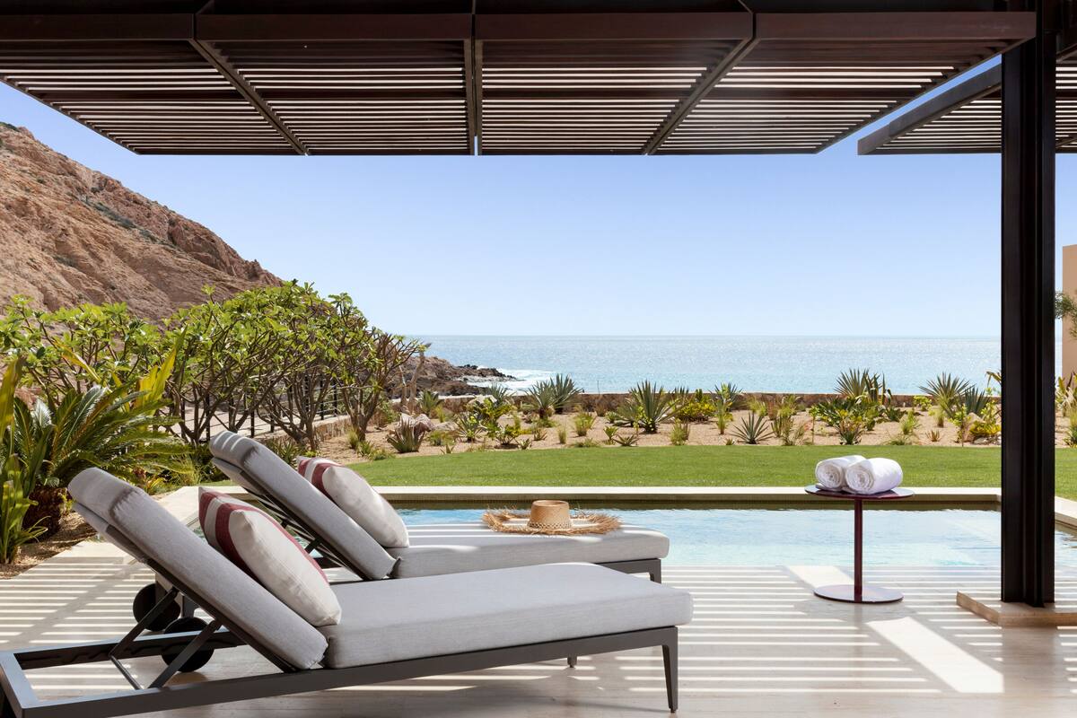 A poolside area with lounge chairs, a small table with towels, and a view of the ocean and a rocky hillside.