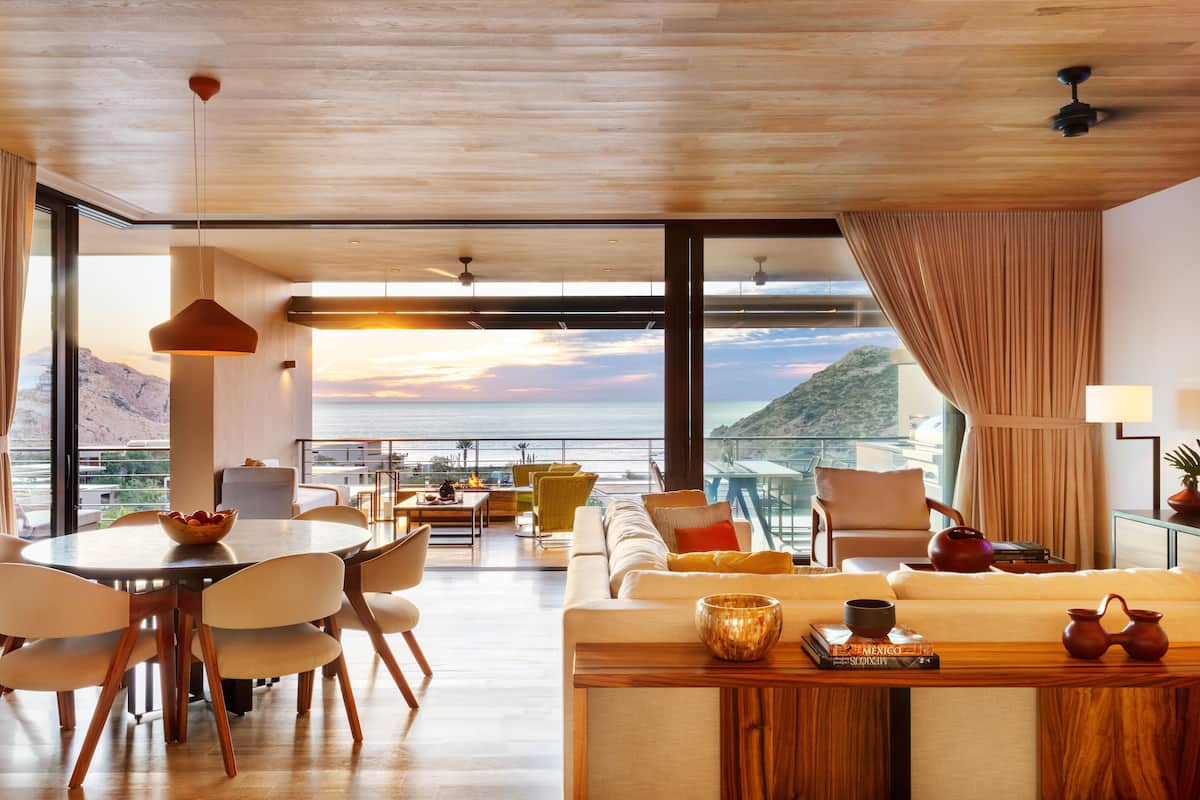 A spacious living area with a large window overlooking a scenic view, a dining area with a round table, and a cozy seating area with a sofa and armchairs.